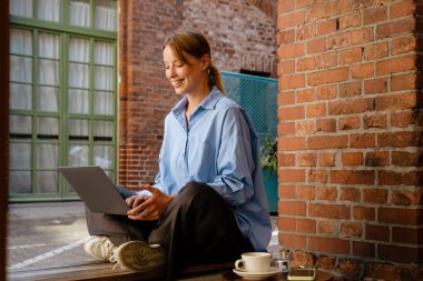 White young woman smiling and using laptop while resting in cafe outdoors