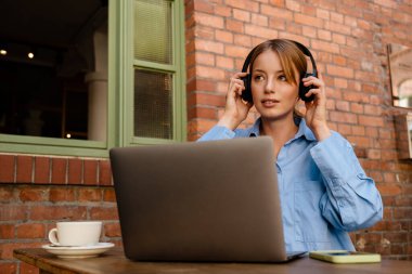 Ginger white woman in headphones working with laptop while sitting in cafe indoors