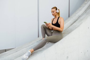 White young woman in headphones using mobile phone after yoga practice outdoors