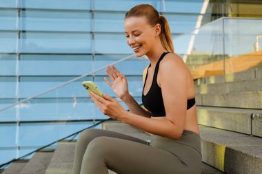 White young blond woman wearing sports bra using cellphone while sitting on stairs outdoors