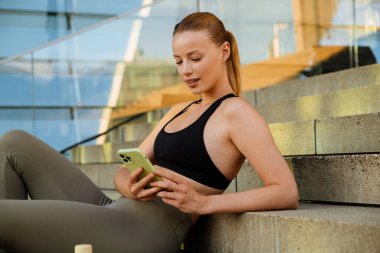 White young blond woman wearing sports bra using cellphone while sitting on stairs outdoors
