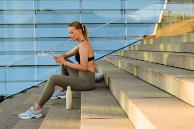 White young blond woman wearing sports bra using cellphone while sitting on stairs outdoors
