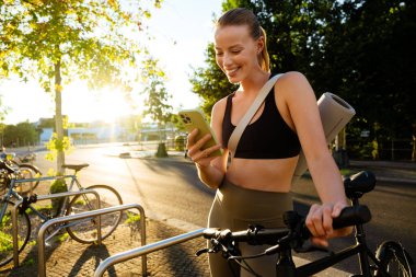 White young woman using cellphone and standing by bicycle after workout outdoors
