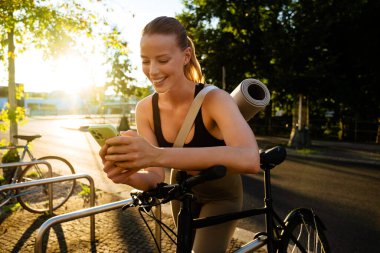 White young woman using cellphone and standing by bicycle after workout outdoors