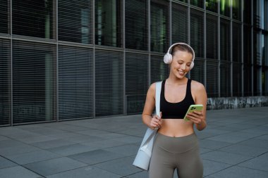 White young woman wearing headphones using cellphone while walking after workout outdoors