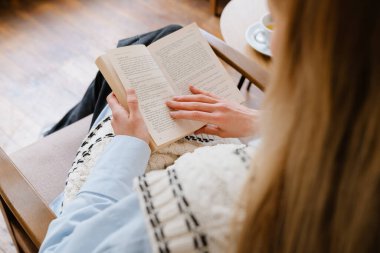 Blonde white woman reading book while sitting in armchair at cafe indoors