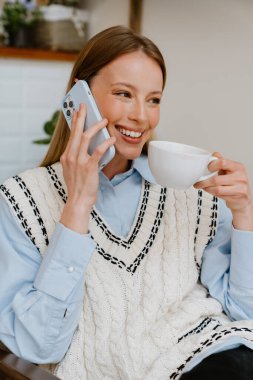 Blonde white woman talking on cellphone while drinking tea at cafe indoors