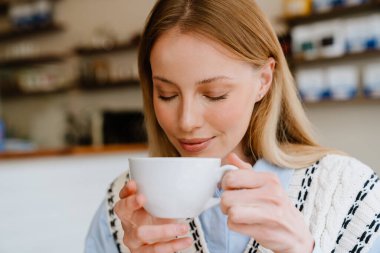 Blonde white woman smiling and drinking tea while sitting at cafe indoors