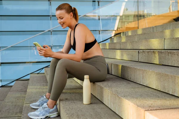 White young blond woman wearing sports bra using cellphone while sitting on stairs outdoors