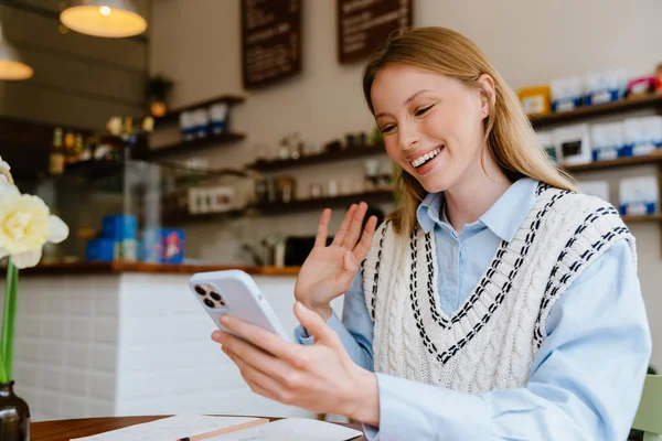Blonde white woman waving hand and using mobile phone while sitting at cafe indoors