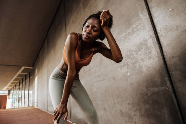 Young sporty tired african woman in headphones wiping her forehead with closed eyes , while leaning on her knees standing indoors