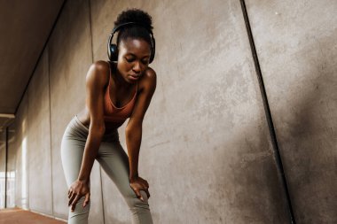 Young sporty calm african woman in headphones looking downward , while leaning on her knees standing indoors