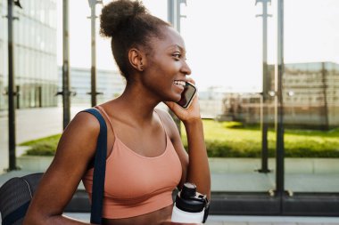 Young beautiful smiling fit afro woman talking phone holding bottle and looking aside standing outdoors