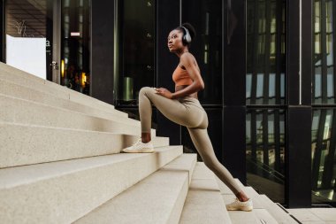 Young beautiful calm african woman in headphones stretching her leg and looking aside, while standing on stairs, near modern building