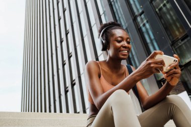 Young beautiful smiling happy african woman in headphones , holding and looking on her phone, while sitting on stairs outdoors