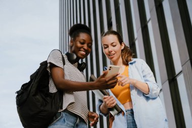 Two young multinational girls smiling and using cellphone together while walking outdoors