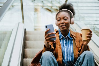Young african american woman using cellphone and drinking coffee while sitting on escalator stairs