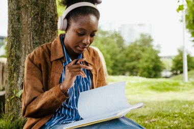 Young beautiful calm african woman in headphones doing homework while sitting in park