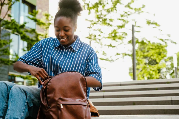 Young Beautiful Smiling African Woman Rummaging Backpack While Sitting Stairs — Stok fotoğraf