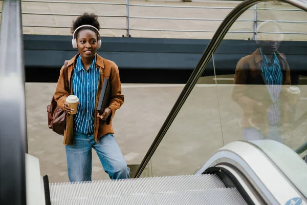 若いです美しいですアフリカの女性でヘッドフォンでコーヒーとフォルダ脇を見ながら 屋外エスカレーターの近くに立って — ストック写真