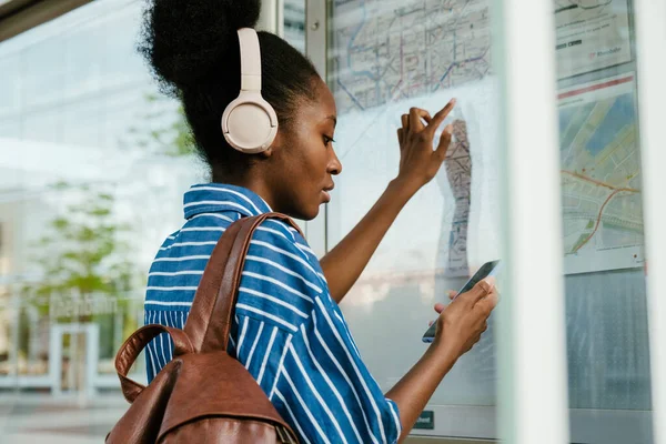 Young african woman in headphones with backpack, pointing on train map considering the route and looking her phone