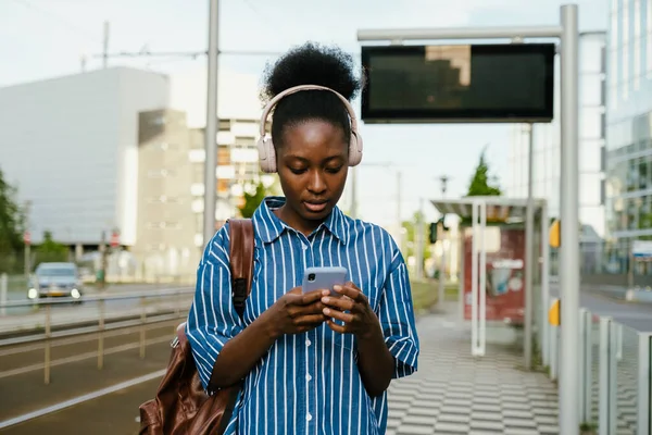 Young african woman in headphones with phone and backpack standing on train station