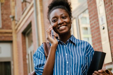 Young beautiful smiling african woman talking phone looking at camera while standing with folder outdoors
