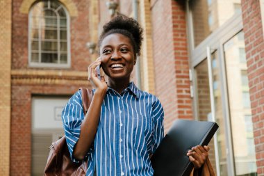 Young beautiful smiling african woman talking phone looking aside while standing with folder outdoors