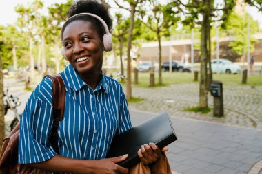 Young beautiful stylish smiling african woman in wireless headphones with book looking aside while standing outdoors