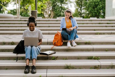 Young multiracial women in headphones doing homework while sitting on stairs outdoors