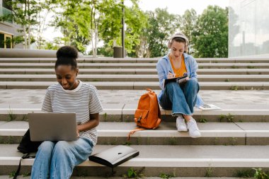 Young multiracial student women doing homework while sitting on stairs outdoors