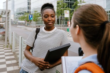 Young multiracial student women talking while standing at city street outdoors