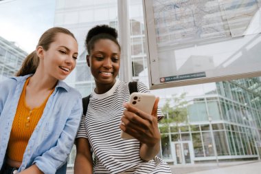 Young multiracial women using mobile phone together while sitting at bus station