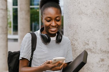 Young black woman smiling and using cellphone while standing at city street