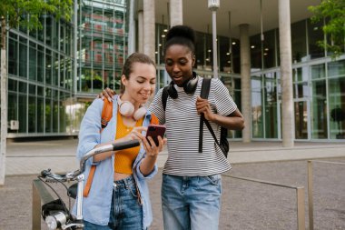Young multiracial women smiling and using cellphone while standing at city street