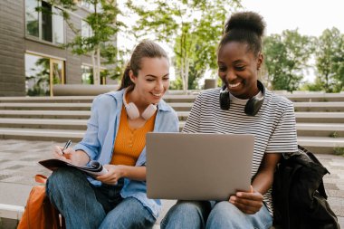 Young multiracial women doing homework with laptop while sitting on stairs outdoors