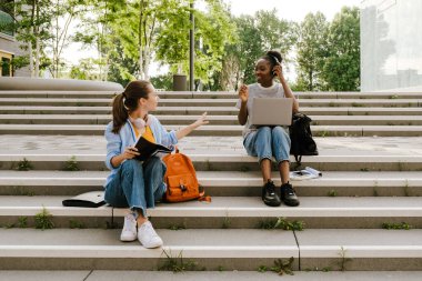 Young multiracial women talking and doing homework while sitting on stairs outdoors