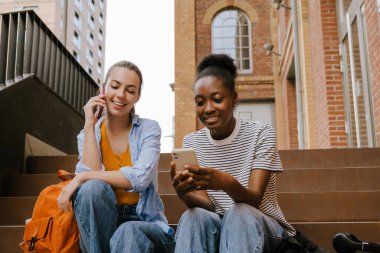 Young multinational girls smiling and using cellphones together while sitting on stairs outdoors