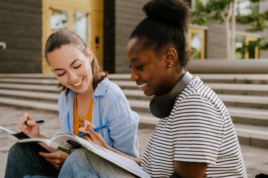 Young multinational girls talking and studying together while sitting on stairs outdoors