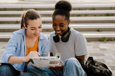 Young multiracial women smiling and doing homework together while sitting on stair outdoors