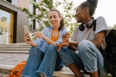 Young multinational girls talking and using cellphone together while sitting on stairs outdoors