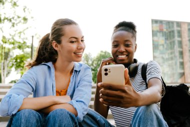 Young multinational girls talking and using cellphone together while sitting on stairs outdoors