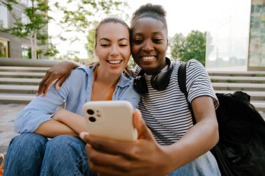 Young multinational girls taking selfie photo on cellphone together while sitting on stairs outdoors