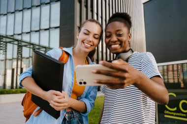 Young multiracial women smiling and taking selfie photo on cellphone at city street