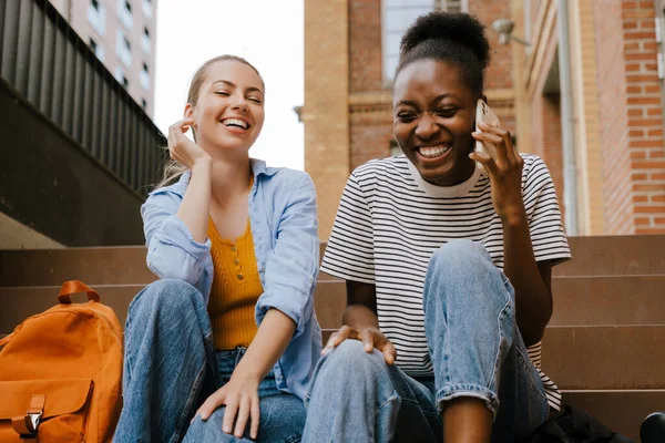 Young Multinational Girls Laughing Using Cellphone Together While Sitting Stairs — Foto Stock