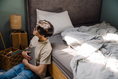 Asian teenage guy holding laptop while sitting on floor at home
