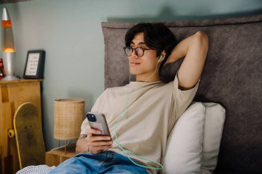 Young asian man in earphones using mobile phone while resting on bed at home