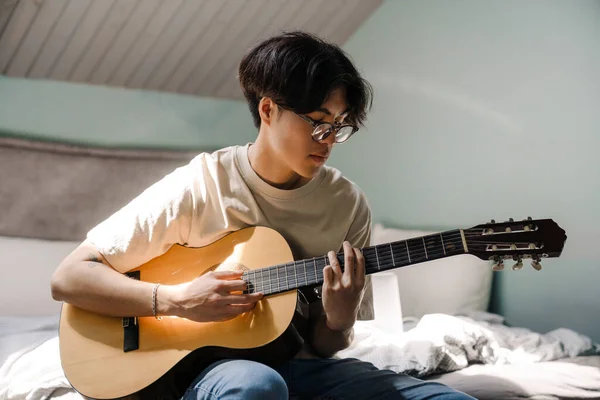 Asian teenage guy playing acoustic guitar while sitting on bed at home