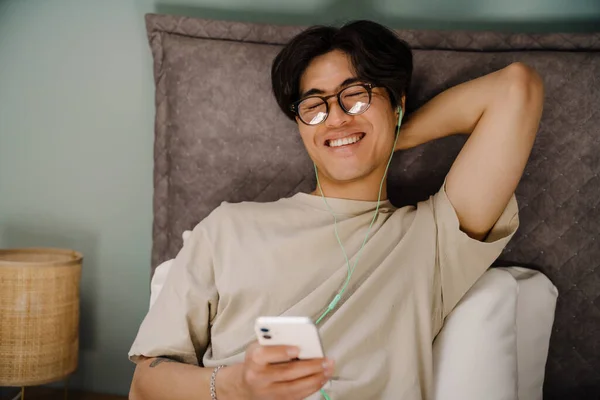 Young asian man in earphones using mobile phone while resting on bed at home