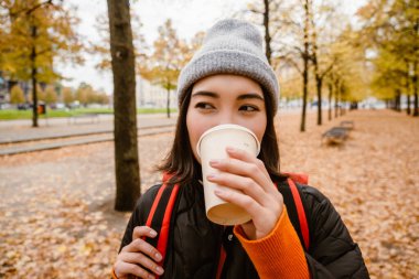 Beautiful young asian woman wearing warm clothes drinking takeaway coffee during walk in autumn park